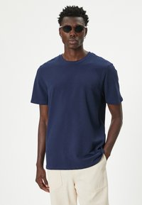 Camiseta de algodón azul marino con cuello redondo y mangas cortas. La tela parece suave y ligeramente texturizada. Se lleva con pantalones de color claro y gafas de sol.