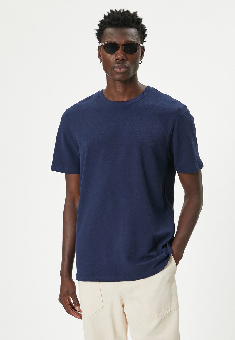 Camiseta de algodón azul marino con cuello redondo y mangas cortas. La tela parece suave y ligeramente texturizada. Se lleva con pantalones de color claro y gafas de sol.