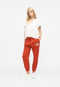 Νεαρή γυναίκα που στέκεται με τα χέρια στις τσέπες, φορώντας λευκό μπλουζάκι με κοντά μανίκια, κόκκινο παντελόνι GAP με καρτούν σχέδιο και λευκά αθλητικά παπούτσια.