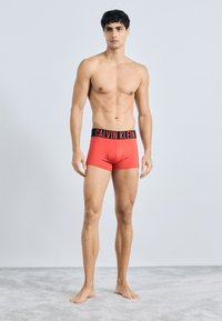 Crvene pamučne bokserice s crnim pojasom na kojem se nalazi logo "CALVIN KLEIN", prilagođenog dizajna, glatke teksture i minimalnih šavova.