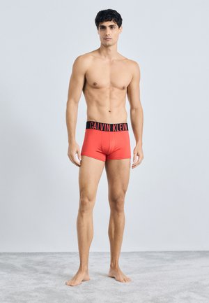 Rde bombažne moške bokserice z črnim pasom, na katerem je logo "CALVIN KLEIN". Imajo prilegajočo se zasnovo, gladko teksturo in minimalne šive.