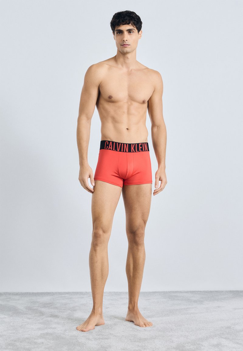 Crvene pamučne bokserice s crnim pojasom na kojem se nalazi logo "CALVIN KLEIN", prilagođenog dizajna, glatke teksture i minimalnih šavova.