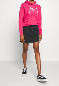 Růžová mikina s kapsou na přední straně a bílým logem, v kombinaci s černou denimovou sukní s knoflíkovým zapínáním a roztrhaným lemováním, doplněná černými teniskami.