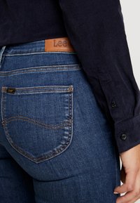 Jeans i denim i mörkblått med en slät yta. Funktioner inkluderar en brun läderetikett i midjan och kontrasterande sömmar på fickorna.