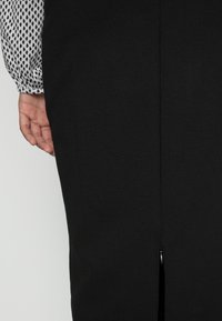 Zwarte jurk met een aansluitend ontwerp, voorzien van een split aan de achterkant; gecombineerd met een blouse met lange mouwen in een zwart-wit ruitpatroon.