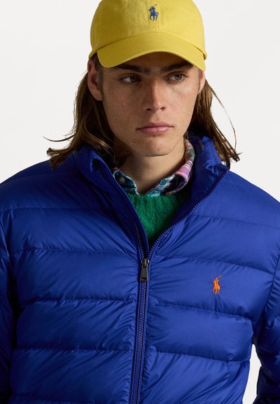 Blå pufferjakke med lynlås foran, orange logo på brystet, båret over en grøn sweater og lagdelt med en ternet skjorte. Gul kasket på hovedet.