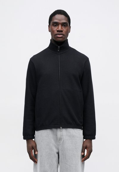 Čierna bundá na zips z textúrovanej látky, s vysokým golierom, dlhými rukávmi a bočnými vreckami, v kombinácii so svetlosivými nohavicami.