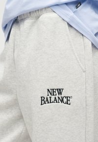 Γκρι φόρμα από μαλακό ύφασμα, με πλαϊνές τσέπες και κεντημένο λογότυπο "NEW BALANCE" σε μαύρο χρώμα στο αριστερό μηρό.