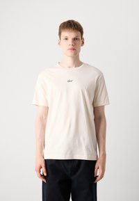 Kortermet, lys beige t-skjorte laget av myk bomull. Har en liten svart logo på brystet. Rett snitt, minimalistisk design.