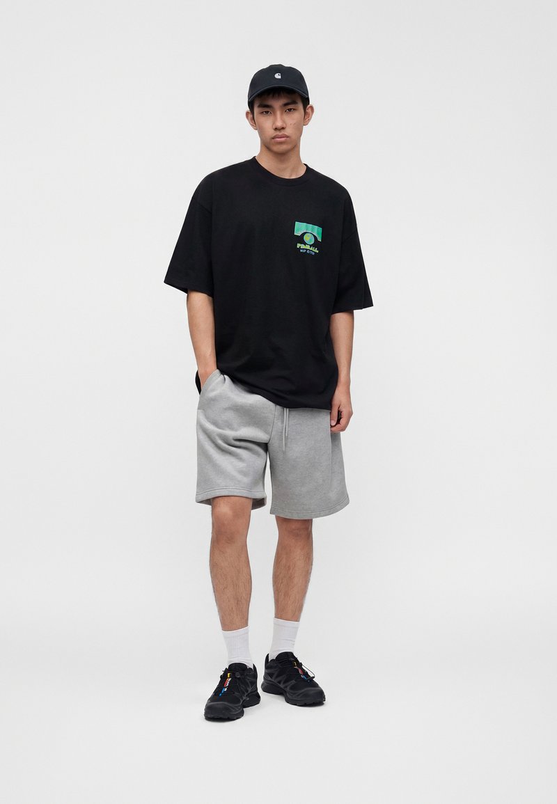 Giovane uomo con t-shirt nera con motivo grafico, pantaloncini grigi, scarpe da ginnastica nere, calzini bianchi e cappellino nero, in piedi davanti a uno sfondo bianco uniforme.