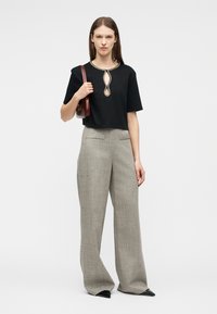 Mujer con blusa negra de manga corta y escote con abertura adornada con cuentas, pantalones grises de pierna ancha, zapatos negros puntiagudos y bolso de hombro granate.