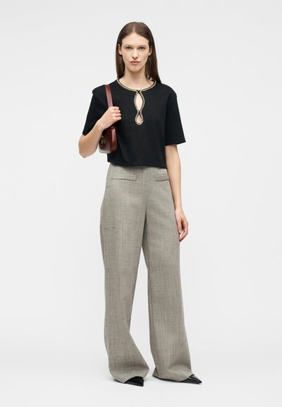 Γυναίκα με μαύρη μπλούζα κοντομάνικη με λαιμόκοψη στολιδωμένη με χάντρες, γκρι παντελόνι φαρδύ, μαύρα μυτερά παπούτσια και κρατάει μπορντό τσάντα ώμου.