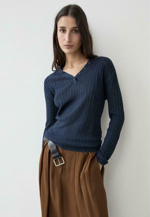 Giovane donna con lunghi capelli scuri che indossa un maglione blu navy a coste con scollo a V e pantaloni marroni larghi con una cintura nera, in piedi contro una parete bianca.