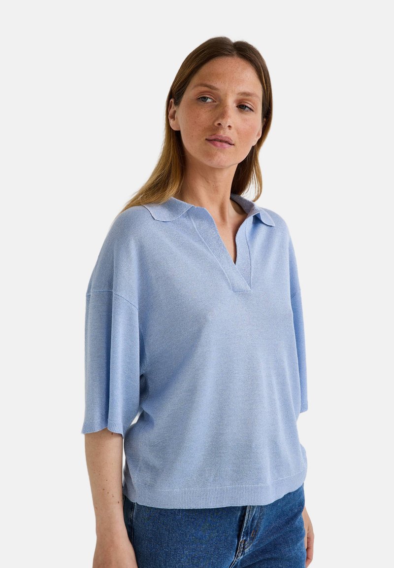 Pull bleu clair avec col, manches courtes et coupe décontractée. Tissu texturé, comportant des détails subtils près du col.