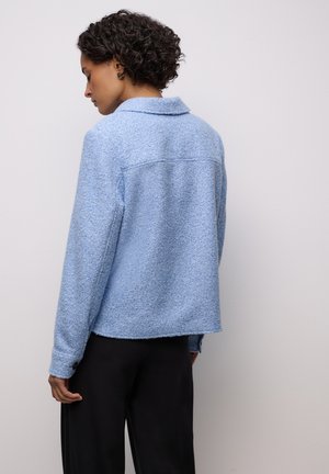 Hellblaue, strukturierte Jacke mit langen Ärmeln, Kragen und Knopfmanschetten, kombiniert mit schwarzen Hosen. Der Stoff wirkt weich und leicht flauschig.