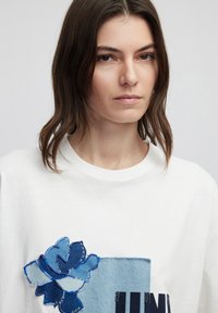 Witte sweatshirt met een blauw bloemenpatch ontwerp in verschillende tinten blauw denim, verlevendigd met gestikte details en een ronde halslijn.