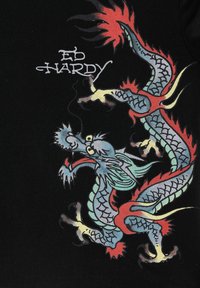 Barevný drak v asijském stylu s modrými šupinami a červenými plameny, umístěný blízko textu "Ed Hardy" na černé tkanině.