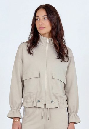 Žena s dlhými tmavými vlasmi, oblečená v béžovej bunde na zips s veľkými prednými vreckami a sťahovacou šnúrkou v páse, pozerajúca sa na stranu.