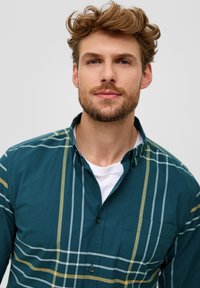 Jonge man met lichtbruin, krullend haar en een baard, gekleed in een groen geruit overhemd boven een wit T-shirt, kijkt recht vooruit.