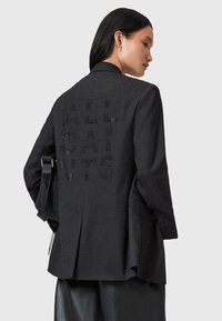Blazer nero con finitura texturizzata, caratterizzato da un grande logo sul retro. Design sartoriale con uno spacco singolo e spalle strutturate.