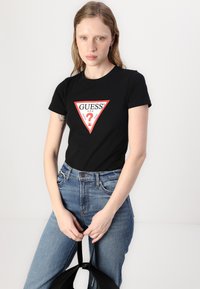 Black cotton t-shirt with a triangular red and white "GUESS" logo on the front, paired with high-waisted blue denim jeans.