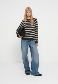 Csíkos, szürke és fekete pulóver, világoskék, bő szabású farmernadrággal párosítva. A modellen egy fekete táska van. Az egyszerű háttér kiemeli az outfitet.