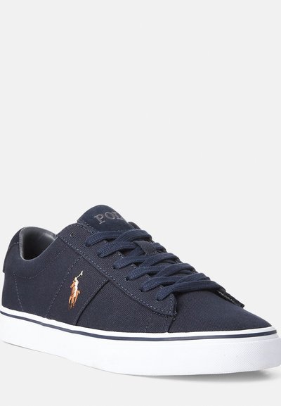 Mørkeblå kanvas sneaker med en hvit gummisåle, flate snorer og en liten brodert logo på siden. Har rund tå og glatt tekstur.