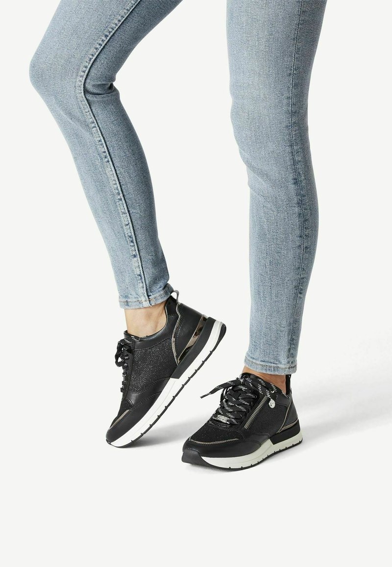 Chaussures de sport noires avec une tige texturée ornée d'accents scintillants et une semelle blanche. Le design comprend des lacets noirs et des fermetures éclair latérales.