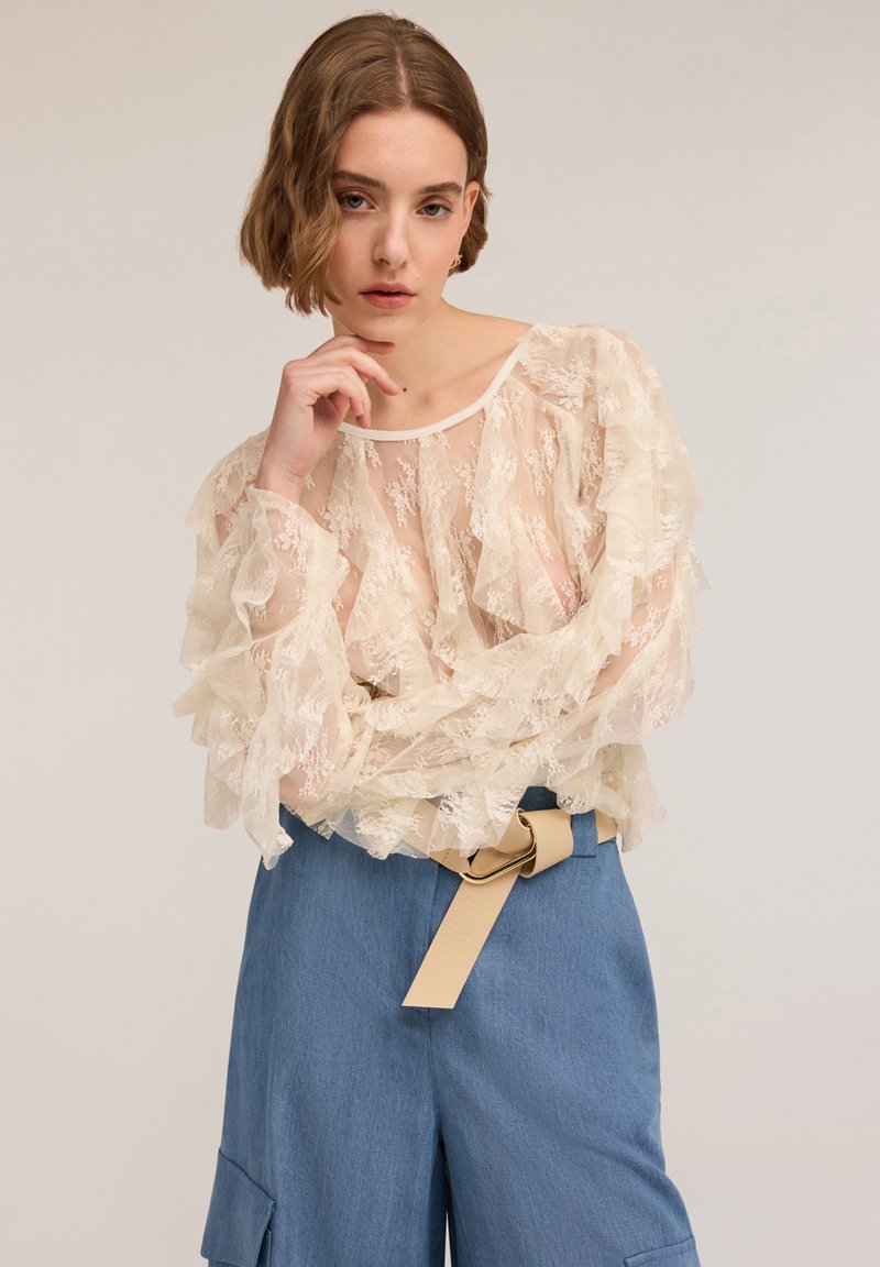 Spitzenbluse mit Rüschendetails in einem cremigen Off-White, kombiniert mit blauen weit geschnittenen Hosen, die einen beigen Gürtel und eine Schnalle aufweisen.