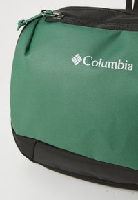 Bolsa verde de Columbia con un tejido texturizado, detalles en negro y cierre de cremallera. Presenta un logo blanco prominente en la parte frontal.