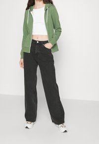 Osoba oblečená v zelené mikině na zip, bílem crop topu, volných černých džínách a bílých teniskách stojí na pozadí bílé stěny.