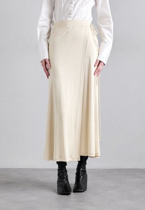 Gonna avvolgente crema realizzata in tessuto liscio; presenta una chiusura con nodo sul lato. Abbinata a stivali neri aderenti e una camicia bianca con bottoni.