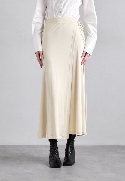Jupe enveloppante crème en tissu lisse ; dotée d'un lien de fermeture sur le côté. Assortie à des bottes noires ajustées et une chemise blanche à boutons.