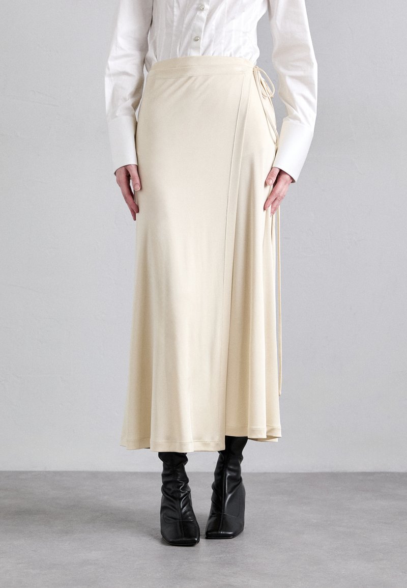 Gonna avvolgente crema realizzata in tessuto liscio; presenta una chiusura con nodo sul lato. Abbinata a stivali neri aderenti e una camicia bianca con bottoni.