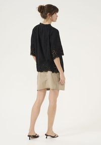 Juoda bluzelė su gėlių akių detalėmis, trumpomis rankovėmis ir susiraukšlėta iškirpte, derinama su smėlio spalvos sijonu ir juodomis aukštakulnėmis sandalais.