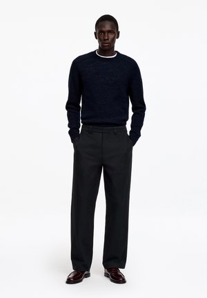 Un homme se tient debout, les mains dans les poches, portant un pull foncé, un pantalon noir à jambes larges et des chaussures marron foncé sur un fond blanc uni.