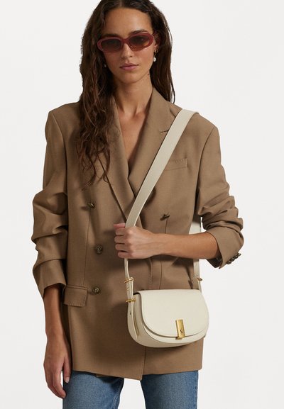 Bežs ādas krustsoma ar izliekta formu, zelta aksesuāriem un regulējamu siksnu. Valkāta ar brūnu žaketi un zilām džinsām.