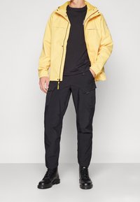 Gul vattentät jacka med huva, svart rundhalsad tröja, svarta cargo-byxor och svarta snörstövlar. Har fickor på jackan och byxorna.