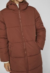 Brūns pufīgs mētelis ar rāvējslēdzēju priekšā, augstu apkakli un regulējamu auklu kapuci. Iekļauj sānu kabatas un quilted faktūru.