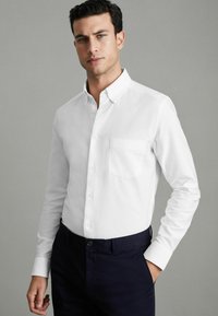 Cămașă albă cu mâneci lungi și un buzunar pe pieptul stâng, purtată cu pantaloni închiși la culoare, are un guler clasic și o textură a materialului neted.