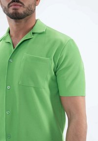 Chemise verte à manches courtes présentant une texture côtelée, un col et une poche sur la poitrine gauche avec des fermetures à bouton.