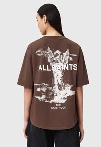 Camiseta oversized marrón con un gráfico de un ángel blanco en la parte posterior que incluye el texto "ALLSAINTS" y "THE SAINTHOOD". Mangas cortas, cuello redondo.