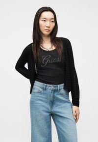 Νεαρή γυναίκα με μακριά σκούρα μαλλιά φοράει μαύρη ζακέτα, μαύρο τοπ «Guess», ασημένιο κολιέ και ψηλόμεσο ανοιχτό μπλε τζιν, στέκεται μπροστά σε λευκό φόντο.