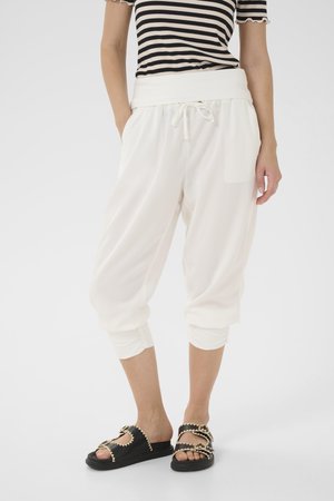 Pantalons plissés blancs à taille élastique avec une texture douce, finitions à poignets froncés et poches, associés à des sandales noires avec un design de bride en zigzag.