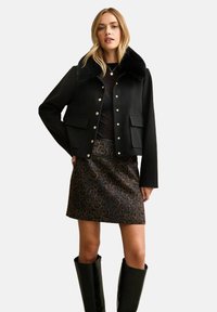 Czarna krótka kurtka z kołnierzem z futerka ekologicznego, zdobiona złotymi guzikami i dwoma przednimi kieszeniami, zestawiona z brązową spódnicą w panterkę i wysokimi kozakami za kolano.
