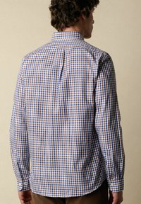 Homme portant une chemise à manches longues à carreaux bleus, bruns et blancs, vu de dos contre un fond beige uni.