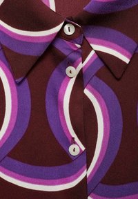 Colletto di camicia con bottoni grandi bianchi su tessuto caratterizzato da audaci strisce curve astratte in viola, rosa e bianco su uno sfondo marrone scuro.