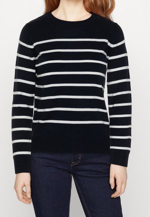 Femme portant un pull noir à manches longues avec des rayures horizontales blanches et un jean bleu foncé, debout devant un fond blanc uni.