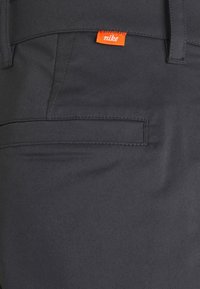 Close-up of dark gray pants back pocket with stitched seam details and small orange Nike label above pocket.