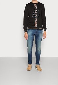 Hombre con sudadera negra con diseño de serpiente, jeans azul desgastados y zapatillas color beige de pie sobre un suelo gris claro contra una pared blanca.